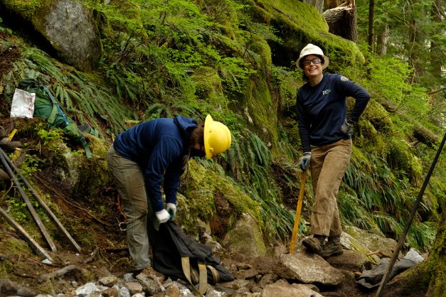 A WCC crew supervisor wearing a white hard hat stands on a rocky trail and smiles at the camera. A crew member in a yellow hard hat bends over to pick up straps.