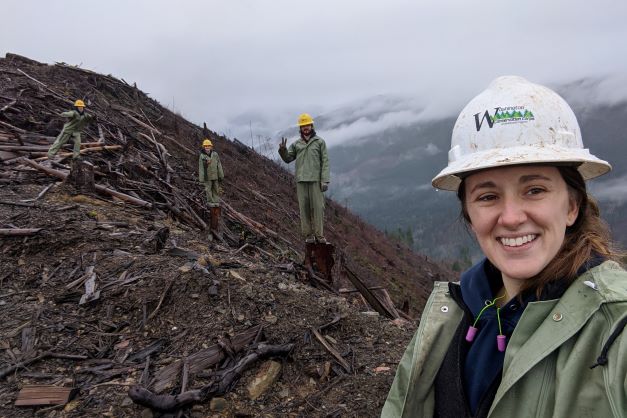 A crew supervisor in a white hard hat smiles in a selfie. Behind them are 3 crew members in rain gear and yellow hats, also smiling. Woody debris along a slope.